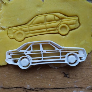 Op de afbeelding: Een witte plastic koekjesvormer in de vorm van een auto. De vormer ligt op een houten oppervlak met gele koekdeeg erachter.