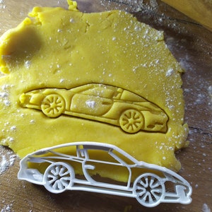 Pode incluir: Um cortador de biscoitos de plástico branco em forma de carro esportivo. O cortador está sobre uma superfície amarela com pó branco sobre ela.