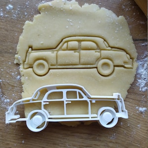 May include: A white plastic cookie cutter in the shape of a vintage car. The cutter is on top of a piece of dough, which is on a wooden surface.