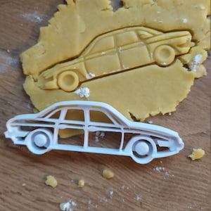 May include: A white plastic cookie cutter shaped like a car. The cutter is on a wooden surface with a piece of cookie dough that has been cut out with the cutter.