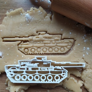 Puede incluir: Un cortador de galletas de plástico blanco con forma de tanque, sobre una superficie de masa marrón espolvoreada con polvo blanco. Un rodillo de madera está en el fondo.