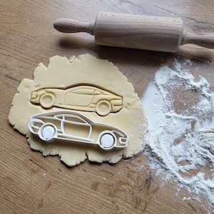 Puede incluir: Un rodillo de madera descansa sobre una superficie de madera junto a un trozo de masa con dos cortadores de galletas con forma de coche. Un cortador está presionado en la masa y el otro está colocado encima. Harina espolvoreada a un lado.