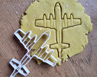 May include: A white plastic cookie cutter shaped like a vintage airplane. The cookie cutter is on top of a piece of raw cookie dough.