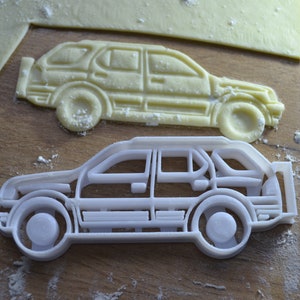 May include: Two white plastic cookie cutters shaped like a car. The cutters are on a wooden surface covered in white flour.