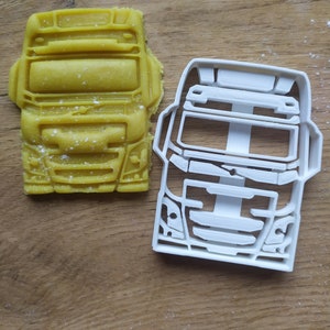 May include: A white plastic cookie cutter shaped like the front of a semi-truck. The cookie cutter is next to a yellow cookie that has been cut out using the cookie cutter.