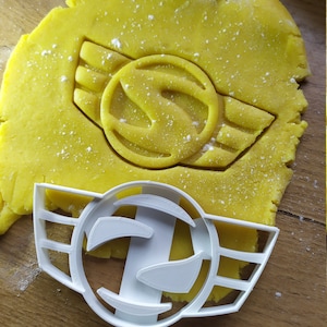 Könnte beinhalten: Ein weißer Kunststoff-Ausstecher in Form eines Logos mit Flügeln, neben einem Ausschnitt in gelbem Teig. Der Teig hat einen passenden Logo-Aufdruck. Das Logo zeigt einen Kreis mit dem Buchstaben "S" im Inneren und Flügeln.