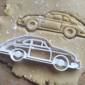 May include: A white plastic car-shaped cookie cutter and a car-shaped cut-out in dough. The cutter is in the shape of a classic car with wheels, windows, and body details. The dough is light brown and dusted with flour, ready for baking.