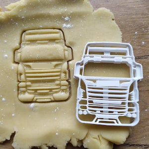 May include: A white plastic cookie cutter shaped like the front of a semi-truck. The cutter is on top of a sheet of unbaked cookie dough.