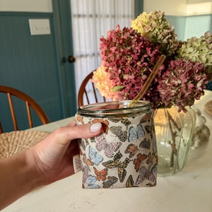 May include: A glass jar featuring a fabric sleeve with a repeating pattern of colorful butterflies in shades of orange, blue, and black. A small white tag labeled Salt & Kin Co. is attached to the side, and a thin amber glass straw rests inside.