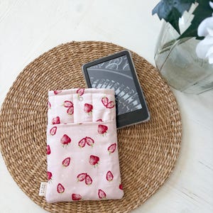May include: A pink ebook reader case with a strawberry pattern. The case is open, revealing a black ebook reader. The case is made of fabric and has a button closure. The case is on a woven mat.