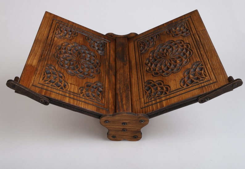 Op de afbeelding: Een houten boekenstandaard met ingewikkelde gesneden ontwerpen aan de voor- en achterkant. De standaard is gemaakt van donkerbruin hout en heeft een scharnierend ontwerp dat het mogelijk maakt om hem te openen en te sluiten.