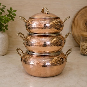 May include: A set of three stacked, round, hammered copper pots with polished metallic surfaces. Each pot features two ornate, textured brass handles on the sides. The top pot includes a matching hammered copper lid with a decorative brass handle.