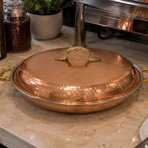 May include: A round, hammered copper serving dish with a matching lid. The dish features ornate, textured gold-toned metal handles on both sides and a matching gold-toned handle on the centre of the lid. The exterior has a polished finish.