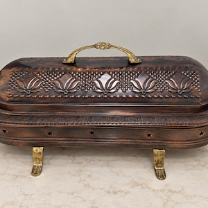 Puede incluir: Una fuente de servir rectangular de color cobre con tapa abovedada que presenta patrones florales y de celosía en relieve. La pieza incluye asas doradas, cuatro patas decorativas y una fila lateral de pequeños recortes en forma de estrella.