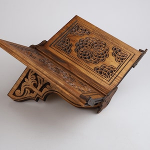 Op de afbeelding: Een houten boekenstandaard met ingewikkelde houtsnijwerk. De standaard is gemaakt van donker hout en heeft een geometrisch ontwerp. De standaard is open en klaar om een boek te houden.
