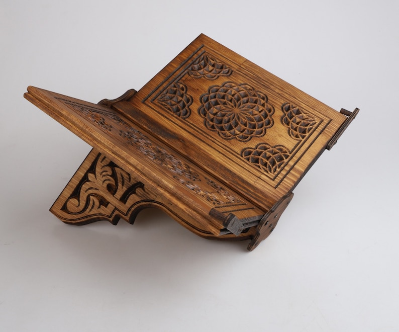 Op de afbeelding: Een houten boekenstandaard met ingewikkelde houtsnijwerk. De standaard is gemaakt van donker hout en heeft een geometrisch ontwerp. De standaard is open en klaar om een boek te houden.