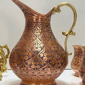 May include: A copper pitcher with an intricate floral design and a brass handle. The pitcher has a round body and a wide mouth.