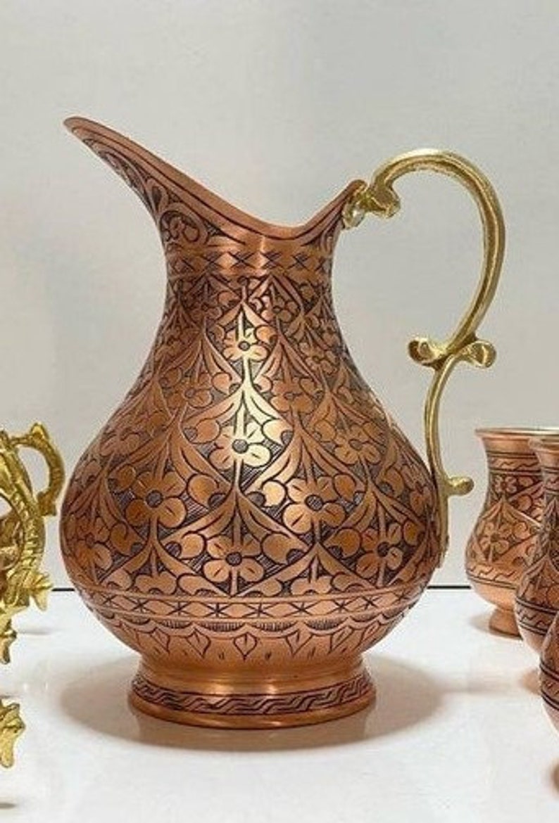 May include: A copper pitcher with an intricate floral design and a brass handle. The pitcher has a round body and a wide mouth.