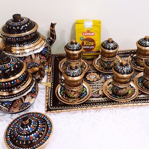 Peut inclure: Un service à thé turc orné, comprenant une théière, des tasses, des soucoupes et un plateau de service. L'ensemble présente des motifs complexes en noir, or et des touches colorées. Un paquet de thé turc est également présent.