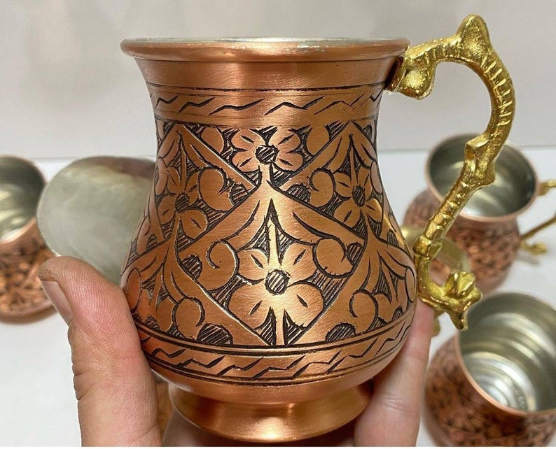May include: Set of four copper mugs with intricate floral designs and a hammered finish. Each mug has a curved handle and a wide base.