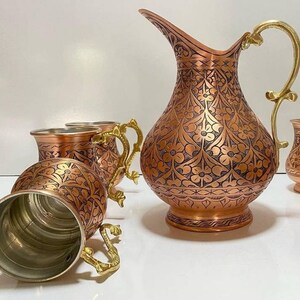 May include: A set of copper mugs and a pitcher with intricate floral designs. The mugs and pitcher have gold-colored handles.