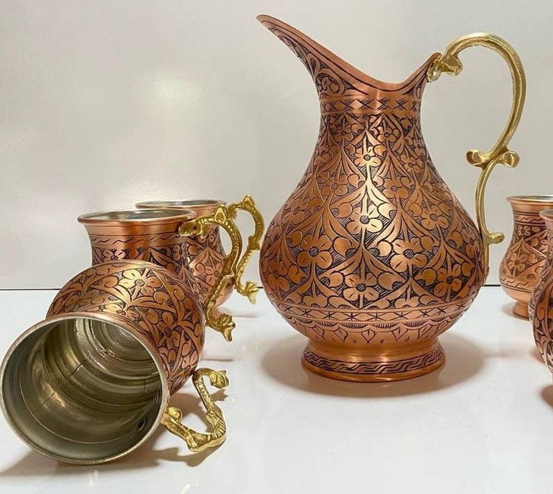 May include: A set of copper mugs and a pitcher with intricate floral designs. The mugs and pitcher have gold-colored handles.