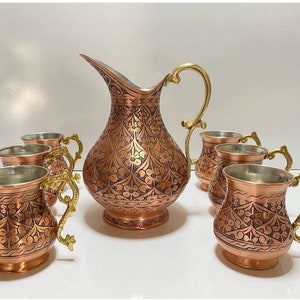 May include: A set of six copper mugs and a copper pitcher with intricate floral designs. The mugs and pitcher have gold-colored handles.