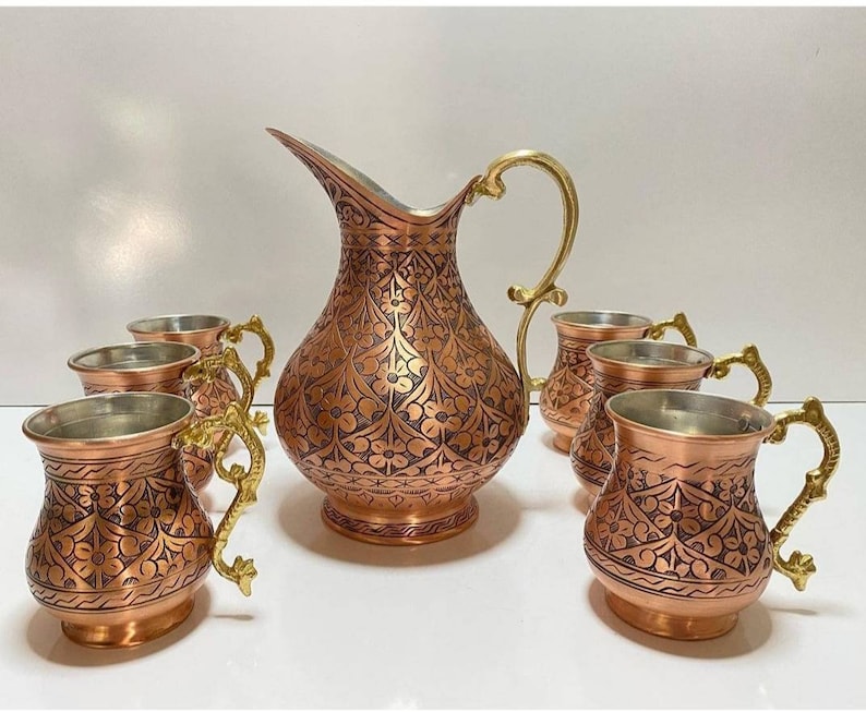 May include: A set of six copper mugs and a copper pitcher with intricate floral designs. The mugs and pitcher have gold-colored handles.