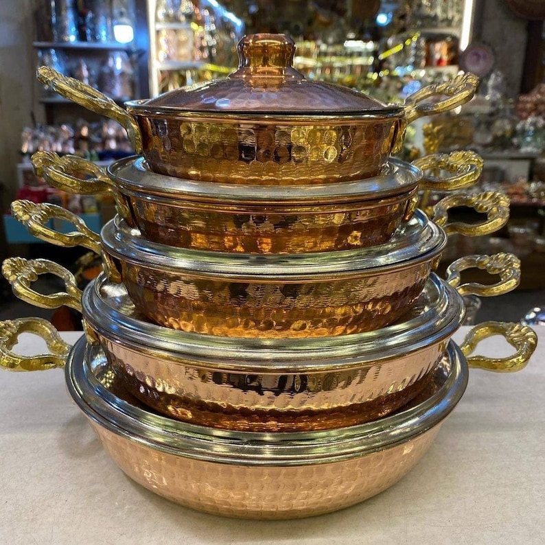 May include: A set of five copper pots with hammered finishes and gold-toned handles. The pots are stacked on top of each other, with the largest pot at the bottom and the smallest pot at the top.