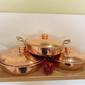 May include: Three copper cookware pieces with hammered surfaces and brass handles. The lids are slightly domed and the pieces are stacked on top of each other.