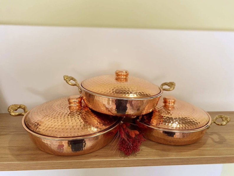 May include: Three copper cookware pieces with hammered surfaces and brass handles. The lids are slightly domed and the pieces are stacked on top of each other.
