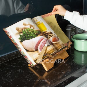 Puede incluir: Un atril de madera para libros de cocina con un marco ajustable que sostiene un libro abierto. La p&aacute;gina muestra el t&iacute;tulo Poultry con im&aacute;genes de carne cruda, hierbas frescas, jengibre y aceite. El soporte tiene un acabado de madera natural.