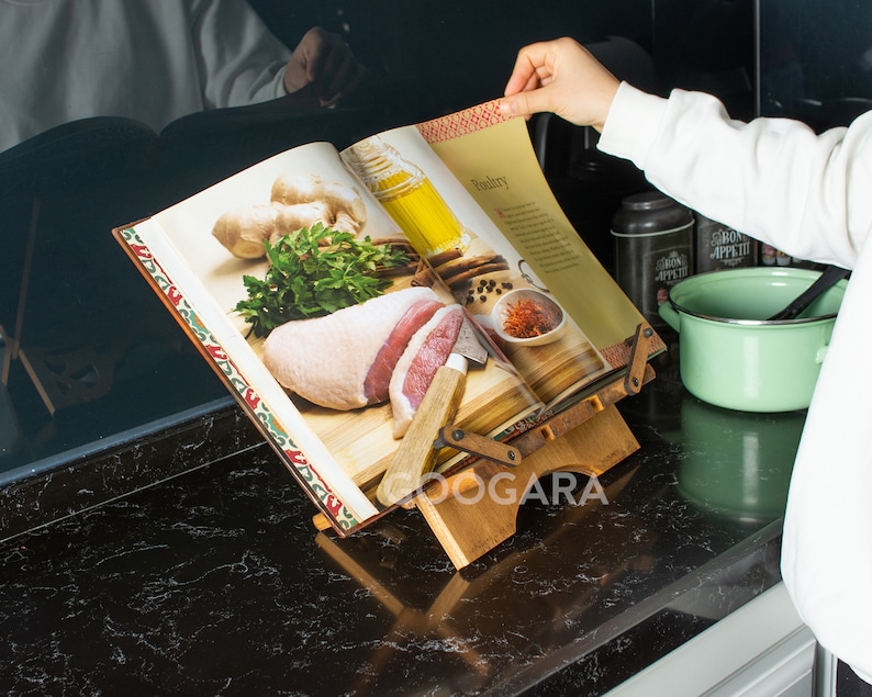 Puede incluir: Un atril de madera para libros de cocina con un marco ajustable que sostiene un libro abierto. La p&aacute;gina muestra el t&iacute;tulo Poultry con im&aacute;genes de carne cruda, hierbas frescas, jengibre y aceite. El soporte tiene un acabado de madera natural.