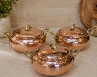 May include: Three round hammered copper pots with matching lids and ornate gold-toned metal handles on each side. The pots feature a textured, dimpled surface and a polished metallic finish, arranged in a cluster on a light-coloured surface.