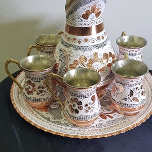 May include: A set of copper mugs and a pitcher with intricate floral designs and gold accents. The set is on a white tray with a gold rim.