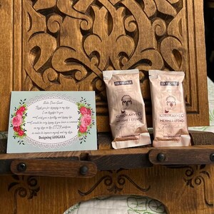 Puede incluir: Dos paquetes de caf&eacute; individuales de color beige met&aacute;lico etiquetados como Kurukahveci Mehmet Efendi T&uuml;rk Kahvesi junto a una tarjeta de agradecimiento rectangular con bordes florales y el texto Designing GOOGARA, sobre un soporte de madera tallada.