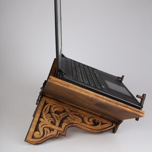 Op de afbeelding: Een houten laptopstandaard met ingewikkelde houtsnijwerk. De standaard is gekanteld om een laptop te ondersteunen, die gesloten is. De standaard is gemaakt van donker hout en heeft een natuurlijke afwerking.