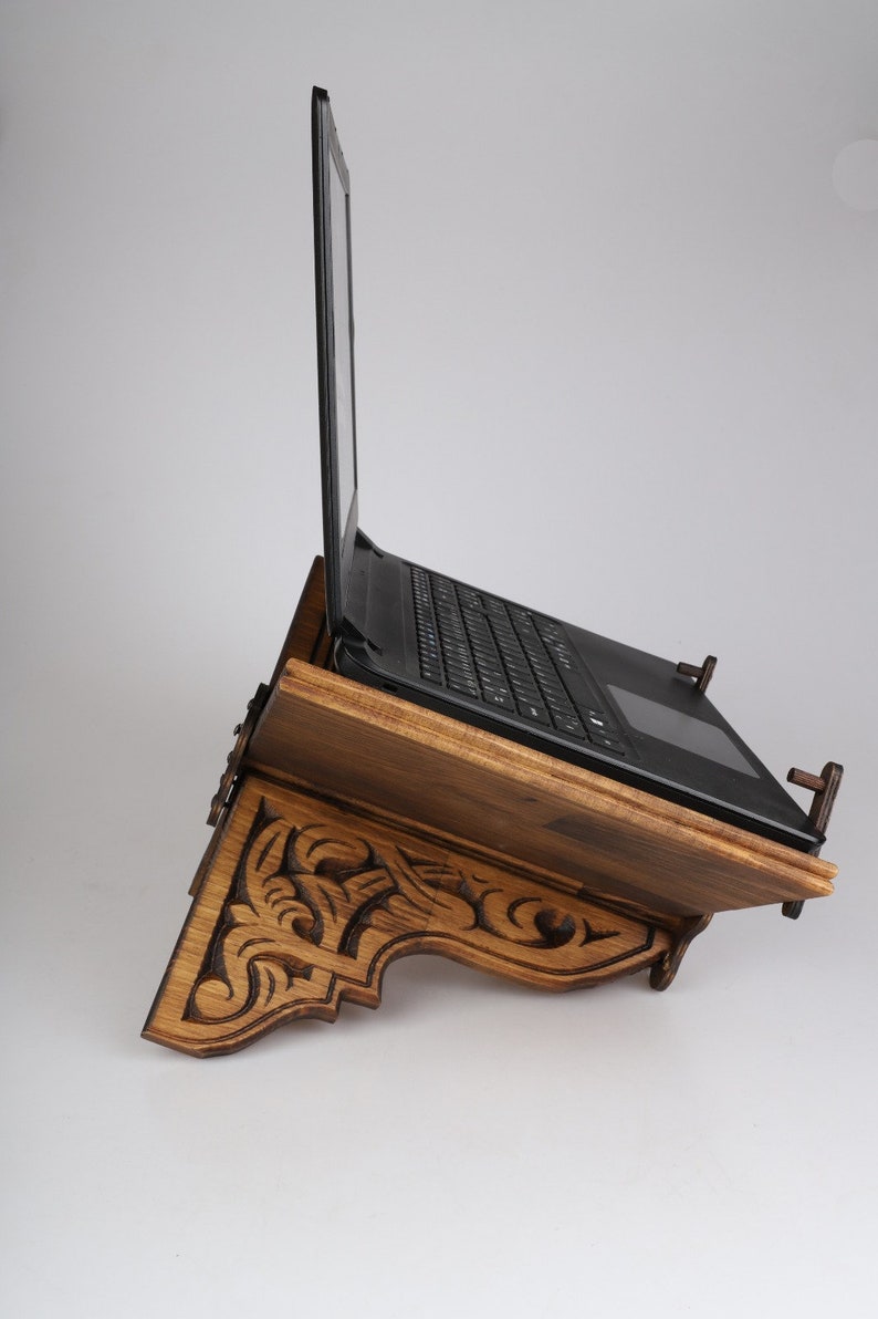 Op de afbeelding: Een houten laptopstandaard met ingewikkelde houtsnijwerk. De standaard is gekanteld om een laptop te ondersteunen, die gesloten is. De standaard is gemaakt van donker hout en heeft een natuurlijke afwerking.