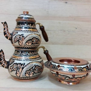 Puede incluir: Un juego de té turco tradicional con una tetera de cobre y un calentador de estufa de cobre a juego. La tetera está decorada con intrincados patrones florales en negro, blanco y dorado. El calentador de estufa tiene un diseño similar.