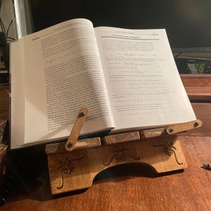 Puede incluir: Un atril de madera con acabado marr&oacute;n oscuro y detalles tallados en volutas sostiene un libro de texto abierto. El libro se titula 6.2 The Feynman propagator y contiene densas ecuaciones matem&aacute;ticas y texto impreso en p&aacute;ginas blancas.