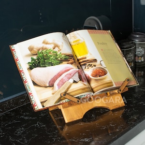 Puede incluir: Un atril de madera para libros de cocina que sostiene un libro abierto titulado Poultry. El soporte presenta un dise&ntilde;o ajustable con dos brazos pivotantes y una base. El libro muestra im&aacute;genes de carne cruda, hierbas, jengibre y especias.