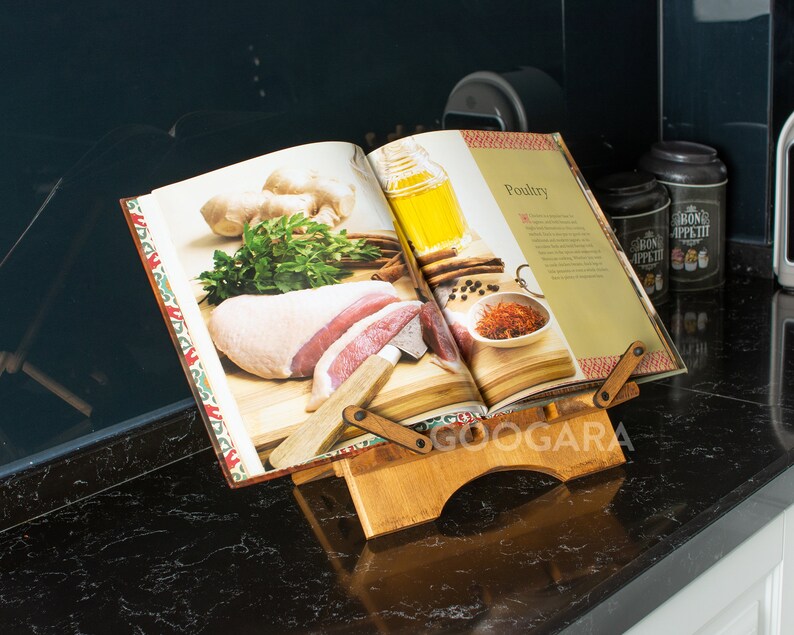 Puede incluir: Un atril de madera para libros de cocina que sostiene un libro abierto titulado Poultry. El soporte presenta un dise&ntilde;o ajustable con dos brazos pivotantes y una base. El libro muestra im&aacute;genes de carne cruda, hierbas, jengibre y especias.