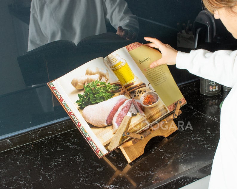 Puede incluir: Un atril de madera para libros de cocina que sostiene un libro abierto en una p&aacute;gina titulada Poultry. El libro muestra una fotograf&iacute;a de carne cruda, jengibre, hierbas y un cuchillo sobre una tabla de cortar. El soporte tiene brazos ajustables.