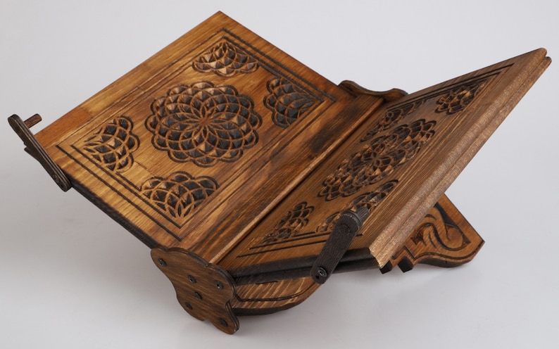 Op de afbeelding: Een houten boekenstandaard met ingewikkelde gesneden ontwerpen. De standaard is gemaakt van donker hout en heeft een scharnierende bovenkant die in verschillende hoeken kan worden aangepast. De standaard is perfect om boeken of andere items te presenteren.