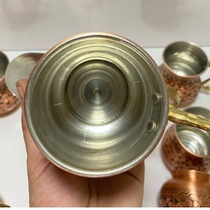 May include: Set of six copper mugs with ornate designs and gold handles. The mugs have a shiny, metallic finish and are perfect for serving beverages.