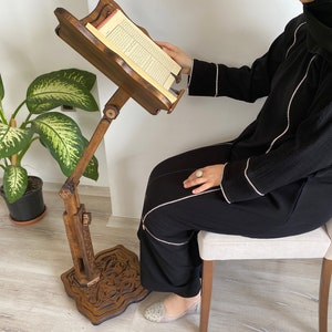 May include: A wooden book stand with intricate carvings on the base. The stand is adjustable and has a book resting on the top. A person is sitting in a chair and reading from the book.