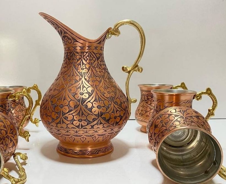 May include: A set of copper mugs and a pitcher with intricate floral designs. The mugs have gold handles and the pitcher has a gold spout and handle. The copper is polished to a shine.
