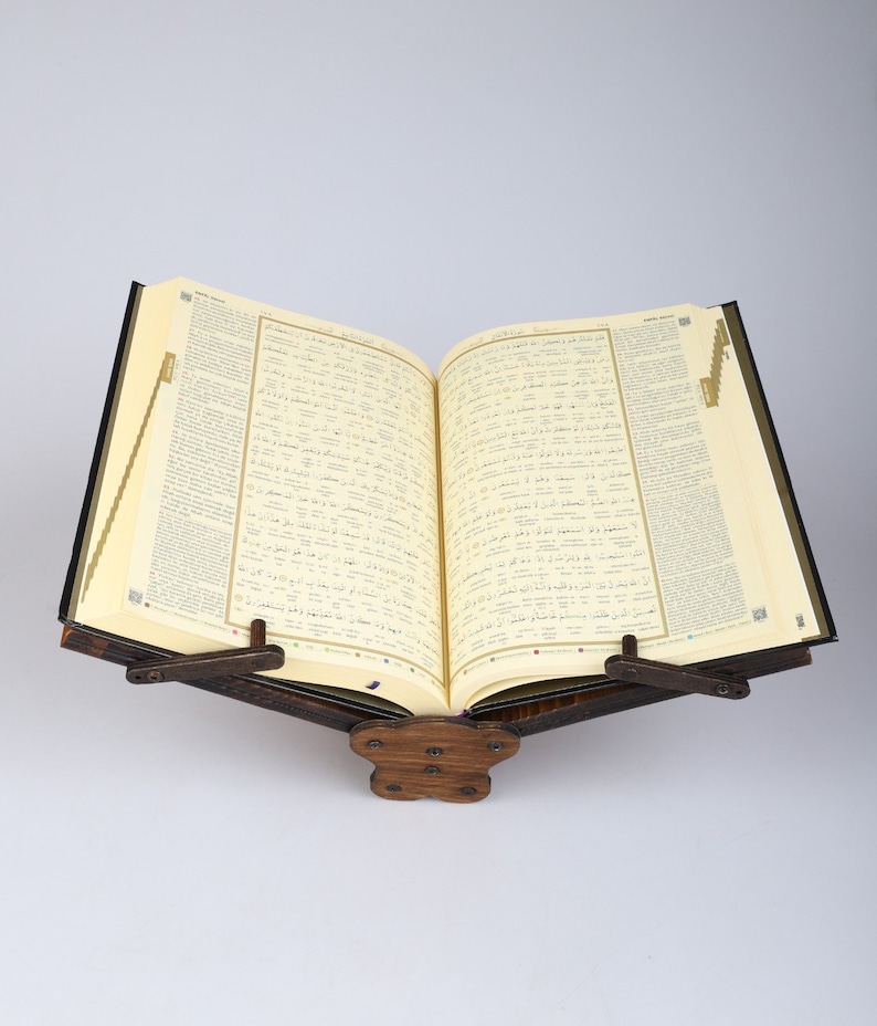 Op de afbeelding: Een houten boekenstandaard met een bruine afwerking, die een groot open boek met Arabische tekst vasthoudt. De boekenstandaard heeft twee metalen scharnieren en een decoratieve basis.