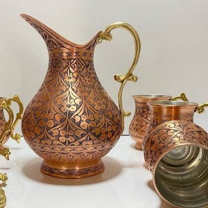 May include: A set of copper mugs and a pitcher with intricate floral designs. The mugs and pitcher have gold-colored handles and are decorated with a dark brown floral pattern.
