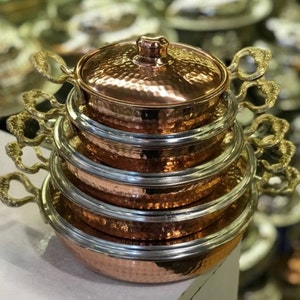 May include: A set of four copper pots with hammered surfaces and brass handles. The pots are stacked on top of each other, with the largest pot at the bottom and the smallest pot at the top. The pots have a shiny, polished finish.
